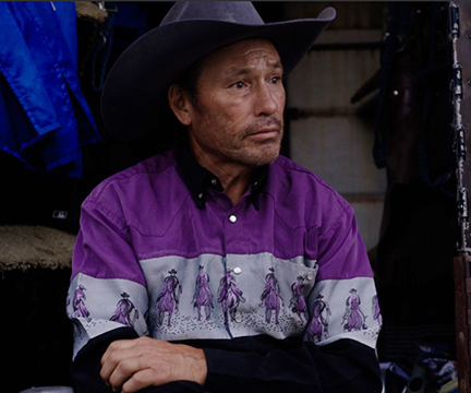 A man in a cowboy hat wearing a purple shit with images on cowboys on horses printed on it looks off to the right. His face is lined and tanned.