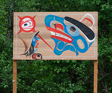 The distinct blue of the Nuxalk Nation seen in a photo on a territory sign features the cultural stylized depiction of a person in a cedar hat looking towards a mythical giant before iit turns into a mountain.