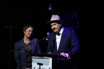 Jasmine Colette looks on as A.W. Cardinal, in a blue suit and grey hat, stands at a mic accepting one of four Maple Blues Awards. 