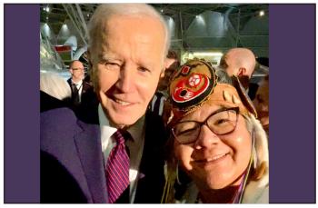President Joe Biden takes a selfie with RoseAnne Archibald. He wears a blue suit and is smiling into the camera. She wears a birchbark headdress with a beaded medallion in the design of the AFN logo at her forehead.