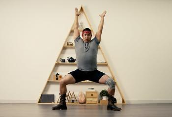 A man has his arms raised high over his head. His legs are wide apart, his knees bent and he’s positioned in a squat as though he is exercising. He poses in front of an A-frame shelf with ornaments on it. He wears a headband, grey T-shirt, shorts and black boots. On one leg he wears an elastic knee brace.
