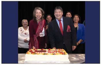 Two people, a woman and a man, stand smiling at the camera before they cut a big cake.