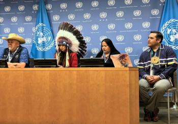 Two men and two women sit at a desk in front of a blue background with the United Nations logo printed on it in white. On man wears a white cowboy hat and one woman wears a feathered chief's bonnet.