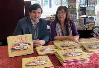 A man and a woman sit behind a table with stacks of books titled The True Canadians.