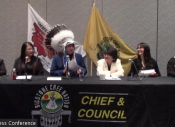 A man wearing a feathered headdress sits at a table and is flanked by others.