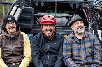 Three men sit on the tailgate of a vehicle laughing togeter. They are wear hats of some kind, but the man in the middle wears a red bike helmet.