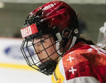 A young woman looks toward the ice. She wears a red hockey helmet with face cage.
