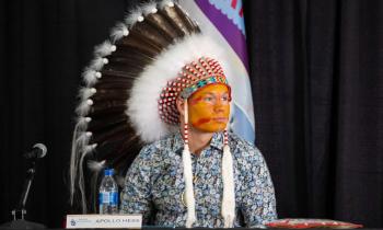 A young man wears a full feather bonnet and ocher-coloured face paint. He sits near a microphone and his name appears on a placecard in front of him--Apollo Hess.  