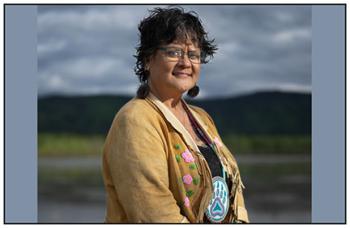 A woman wearing a buckskin jacket and a beaded medallion around her neck stands against a backdrop of a hilly range and cloud-filled sky. 