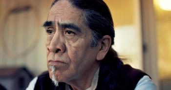 A man with heavy dark eyebrows and a grey patch of a goatee on his chin sits in a room and stares out of frame.