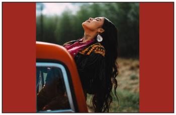 A young woman with long black hair leans backwards out the passenger side of a truck, closes her eyes and raises her face to the sun.