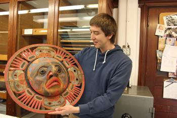 A man stands holding a carved sun mask off to his side.