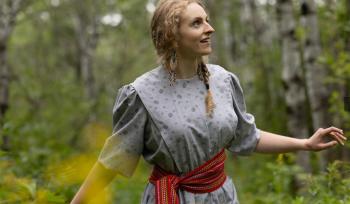 A young girl in a cotton dress with a Metis sash around her waist walks through the woods.