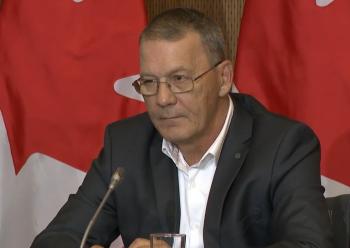 A man sits ibehind a microphone in front of a Canadian flag.