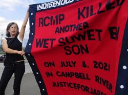Mother of slain man holds up red button blanket with words on it calling out the RCMP for his death.