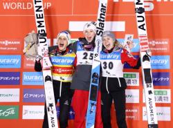 Winners of the women's World Cup ski jumping competition stand in front of an orange wall festooned with multi-coloured plaques with Japanese writing on them. The women hold their skis, bouquets and have their arms around each other. They smile wide with excitement.