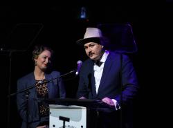 Jasmine Colette looks on as A.W. Cardinal, in a blue suit and grey hat, stands at a mic accepting one of four Maple Blues Awards. 