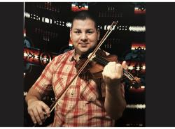 Adam Daigneault wears a red checked short-sleeved shirt. He holds a fiddle under his chin and a bow to the strings. He is standing in front of a blanket with a geometric design. He is starring smiling.