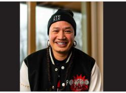 Patrick Mitsuing smiles into the camera. He's wearing a black toque that reads Land Back on it, and a black jacket with white sleeves. It has a logo that reads Powwow.