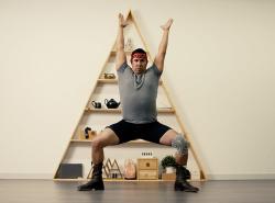 A man has his arms raised high over his head. His legs are wide apart, his knees bent and he’s positioned in a squat as though he is exercising. He poses in front of an A-frame shelf with ornaments on it. He wears a headband, grey T-shirt, shorts and black boots. On one leg he wears an elastic knee brace.