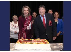 Two people, a woman and a man, stand smiling at the camera before they cut a big cake.