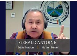 A man is on a livestream call. He wears a buckskin beaded vest and earphones and holds his finger up as he speaks. Behind him is a flag of the Dene Nation. The logo has two rivers. Trees at the bottom, grasslands on eachside and mountains behind. It says "Land of the People."