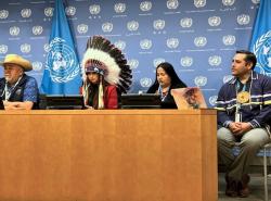 Two men and two women sit at a desk in front of a blue background with the United Nations logo printed on it in white. On man wears a white cowboy hat and one woman wears a feathered chief's bonnet.
