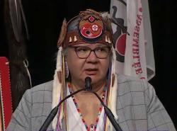A woman in a grey suit speaks at the microphone. She wears a birchbark headband with shells on it and white fur pieces dangling from it. On it is a medallion with the AFN logo on it.