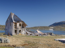 An abandoned wood-constructed home on a grassy and rock-strewn landscape is falling in on itself. The roof is caving in, and one side of house lay on the ground next to a body of water.