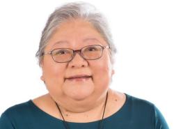 This is a photo of a woman in a teal shirt. She is grey-haired, and wears glasses. She smiles towards the camera. 