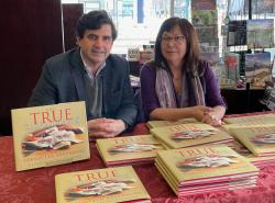 A man and a woman sit behind a table with stacks of books titled The True Canadians.