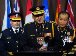 Three police men in full uniform, two wearing hats, stand in front of a bank of flags. One stands before a microphone to speak.