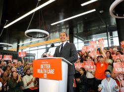 A man stands at a podium surrounded by cheering people displaying NDP election signs.