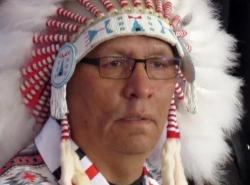 A close-up of the face of a man wearing a feather chief's bonnet is seen.