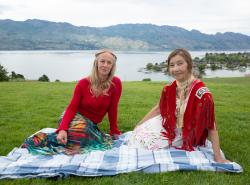 Two women sit on a blue checkered blanket on a green grassy area in front of a lake.