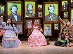 Three women in 1800s garb sit in chairs on a stage in front of framed portraits of men from the time period. The women have their right arms raised above their heads and hands making fists.