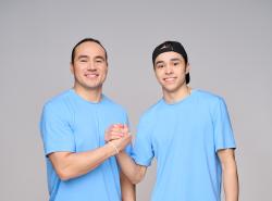A man and his teen son clasp hand and smile towards the camera. They are in matching light blue t-shirts and the teen wears his ball cap backwards on his head.