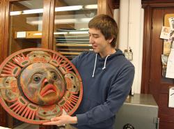 A man stands holding a carved sun mask off to his side.