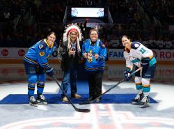A man wearing a feather bonnet and a woman in a hockey jersey stand waiting to drop th puck for two women hockey players.