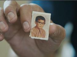 A small school photo of a boy wearing dark-rimmed glasses is held up between the index finger and thumb of a man's hand.