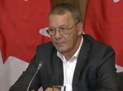A man sits ibehind a microphone in front of a Canadian flag.